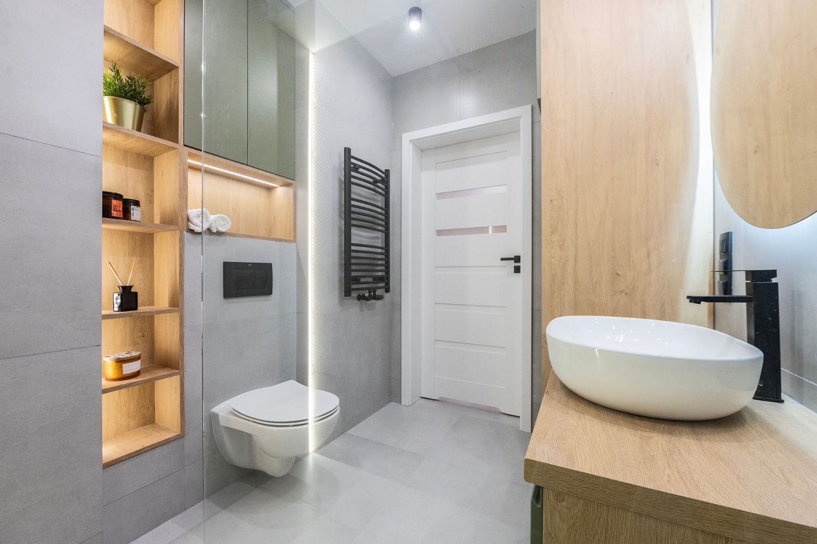 Nowoczesna łazienka z podświetlaną półką, wiszącą toaletą i umywalką nablatową. Szara płytka, drewniane akcenty, minimalistyczny design.