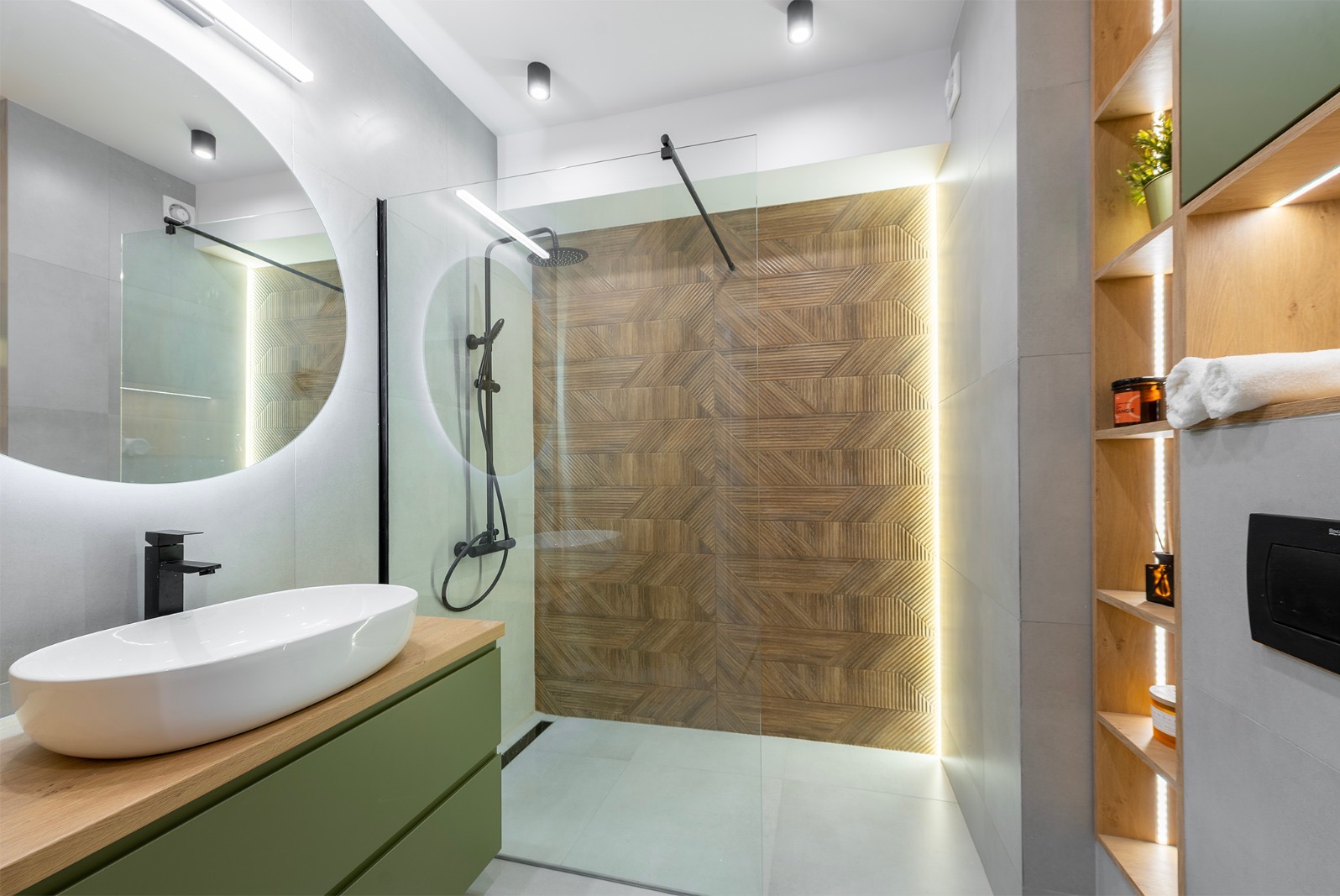 Nowoczesna łazienka z kabiną prysznicową typu walk-in, drewnianą ścianą z podświetleniem LED, okrągłym lustrem i umywalką nablatową na zielonej szafce.