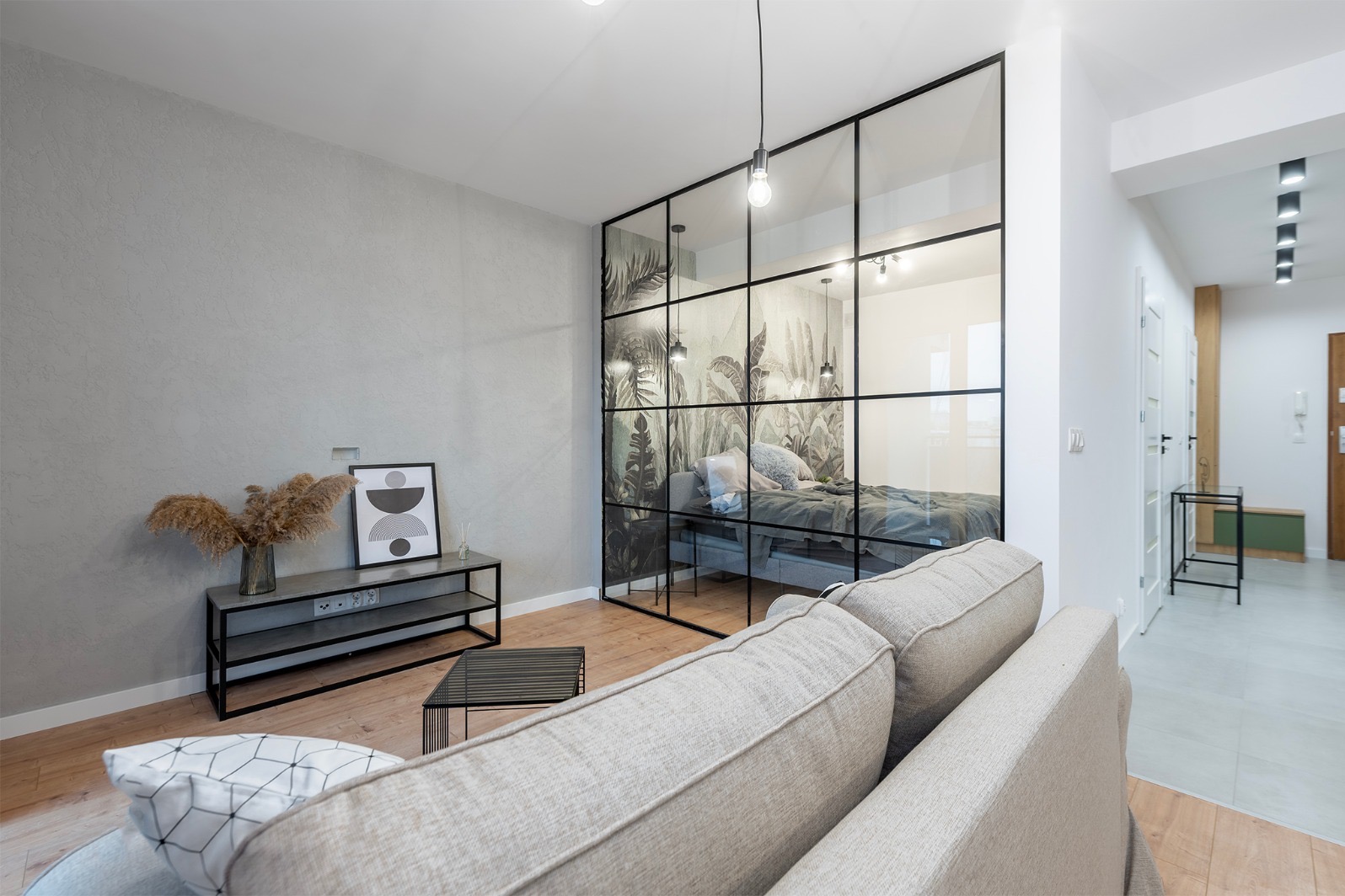 Nowoczesny salon z sofą w neutralnym kolorze, oddzielony szklaną ścianą z motywem roślinnym od sypialni. Minimalistyczny stolik i komoda z dekoracjami.