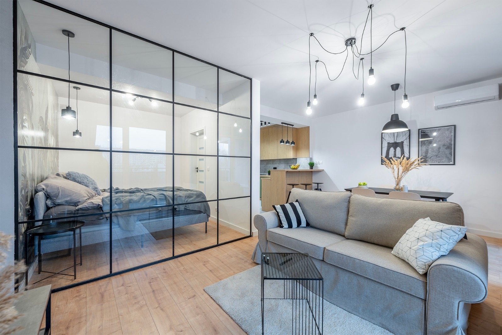 Nowoczesne wnętrze mieszkania z przeszkloną ścianą oddzielającą sypialnię od salonu, drewniana podłoga, szara sofa i designerskie oświetlenie.