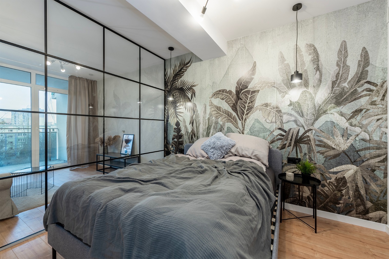 Sypialnia z łóżkiem, ścianą z motywem roślinnym i przeszkleniem w czarnej ramie. Nowoczesny design z akcentami natury i minimalistycznym oświetleniem.