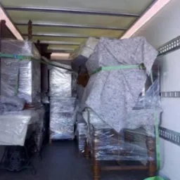 Wnętrze busa wypełnione meblami zabezpieczonymi folią i pasami transportowymi. Widoczna stara maszyna do szycia i komody. Przygotowanie do transportu.