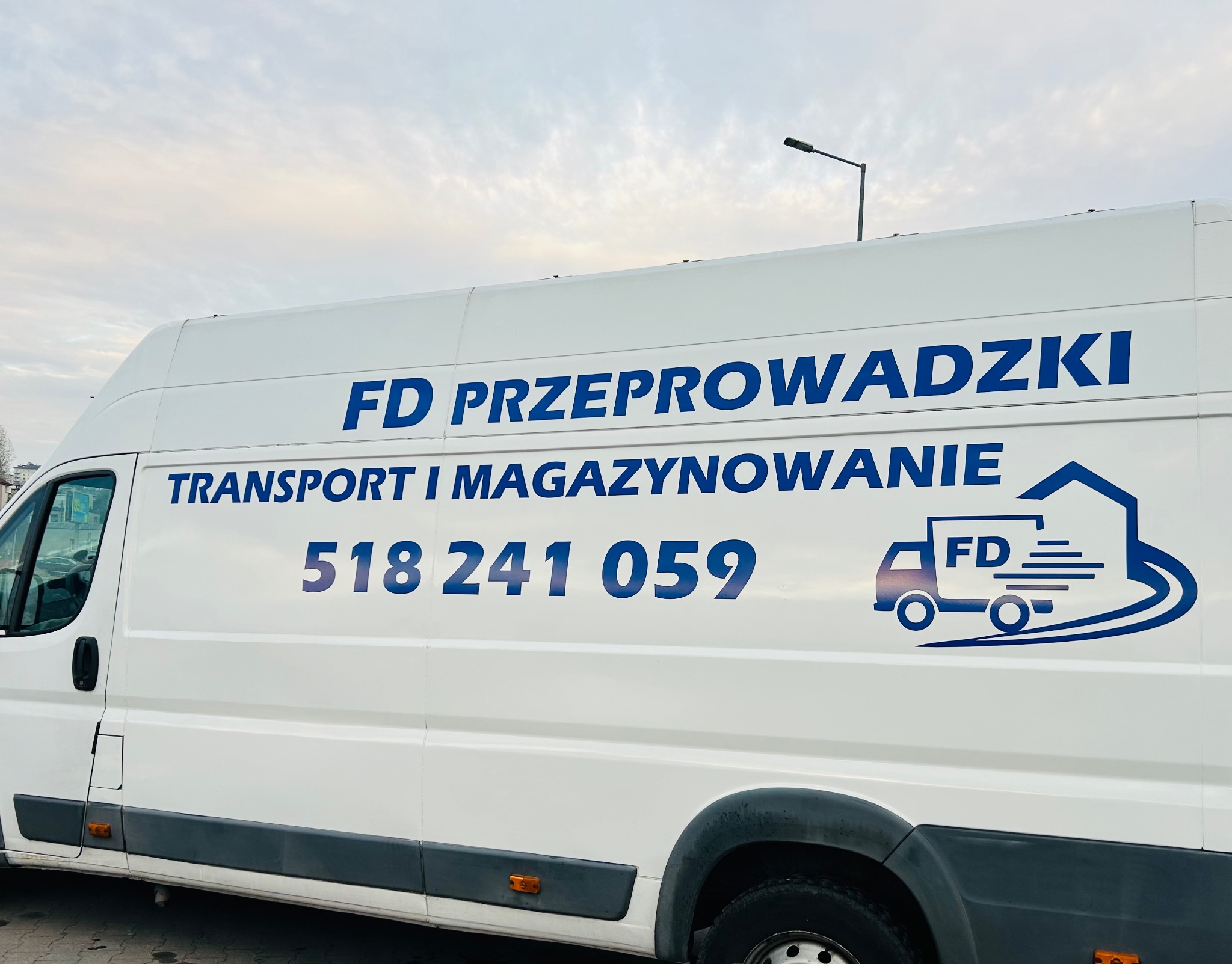 Biały bus z niebieskim logo FD Przeprowadzki, numerem telefonu i grafiką transportu domu. Usługi transportu i magazynowania w tle pochmurne niebo.