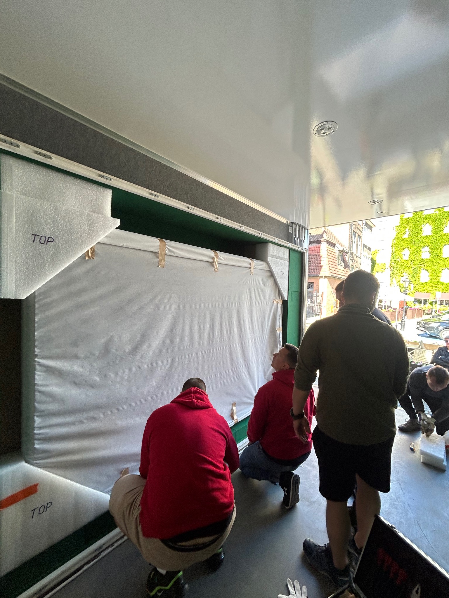 Pracownicy firmy przeprowadzkowej zabezpieczają duży, owinięty w folię obraz wewnątrz pojazdu transportowego. Widoczne oznaczenia 'TOP' na opakowaniu.