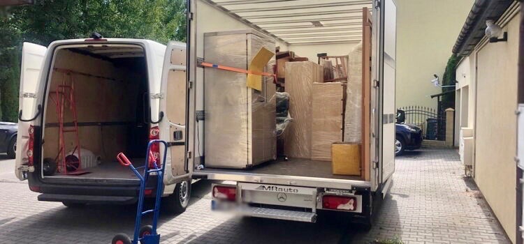 Otwarty bus z meblami i sprzętem AGD zabezpieczonym folią, w tle budynek. W drugim busie wózek transportowy. Przeprowadzka na posesji.