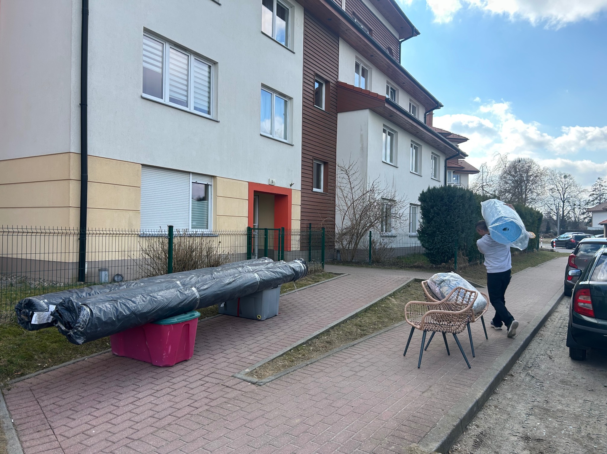 Przed blokiem przygotowania do przeprowadzki: meble ogrodowe, zwinięte dywany w folii, osoba niesie pakunek. Słoneczny dzień.