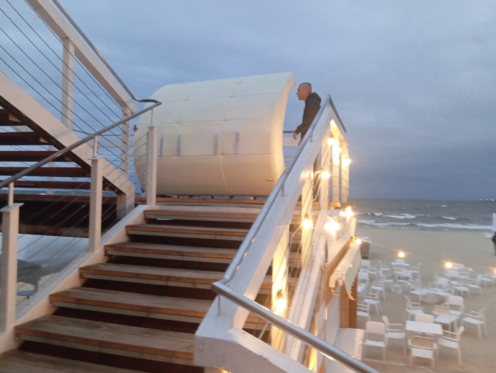 Osoba na schodach z modułową konstrukcją w tle, widok na plażę i morze o zmierzchu, oświetlenie dekoracyjne, białe krzesła i stoły. Nietypowe ustawienie.
