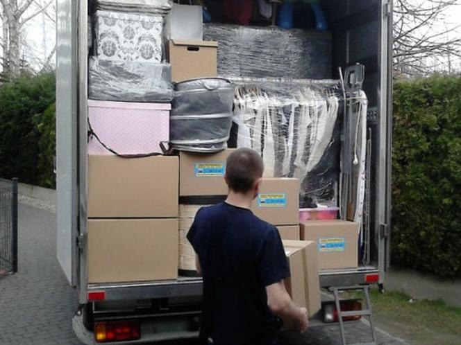 Mężczyzna wnosi karton do wypełnionego po brzegi busa przeprowadzkowego. Widoczne meble, pudła i ubrania zabezpieczone folią stretch. Załadunek w toku.