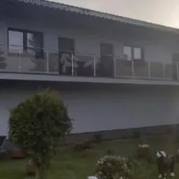 Jasny dom z balkonem i szklanymi barierkami, otoczony zielenią. Widoczny trawnik, krzewy i dekoracje ogrodowe. Ujęcie z dołu, w tle zachodzące słońce.