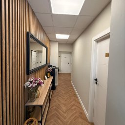 Skóra Balans - Wnętrze salonu z drewnianą ścianą, lustrem i kwiatami. Podłoga w jodełkę, białe drzwi z numerem. Nowoczesny design, jasne oświetlenie i przytulna atmosfera.