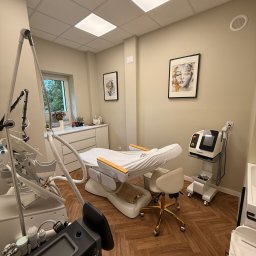 Skóra Balans - Jasne wnętrze gabinetu z leżanką kosmetyczną, krzesłem na złotych kółkach i nowoczesnymi urządzeniami do zabiegów. Na ścianach wiszą obrazy w czarnych ramkach.