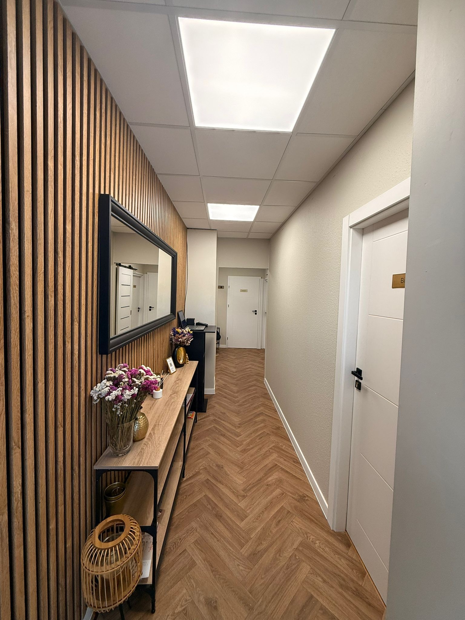 Wnętrze salonu z drewnianą ścianą, lustrem i kwiatami. Podłoga w jodełkę, białe drzwi z numerem. Nowoczesny design, jasne oświetlenie i przytulna atmosfera.