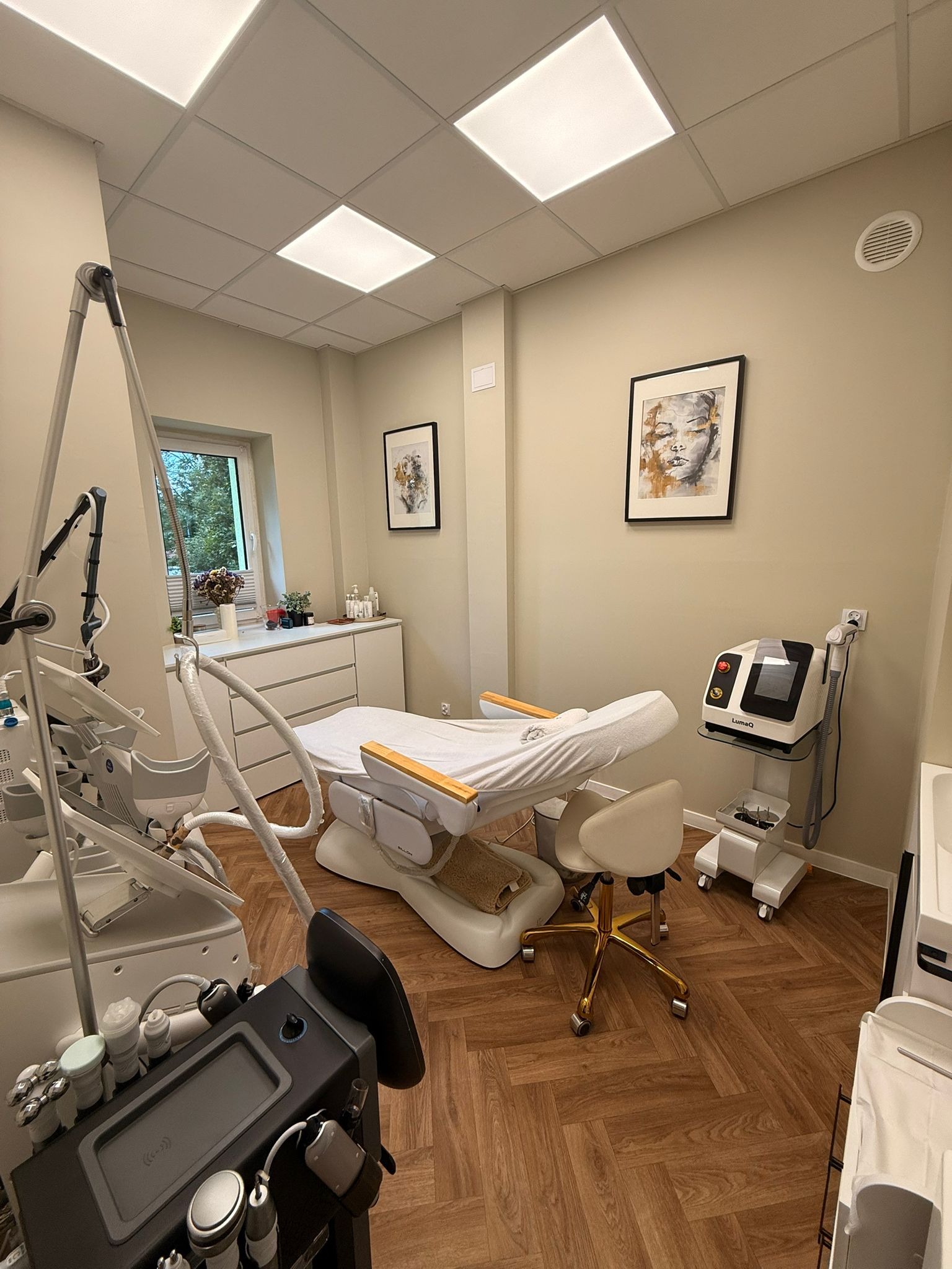 Jasne wnętrze gabinetu z leżanką kosmetyczną, krzesłem na złotych kółkach i nowoczesnymi urządzeniami do zabiegów. Na ścianach wiszą obrazy w czarnych ramkach.