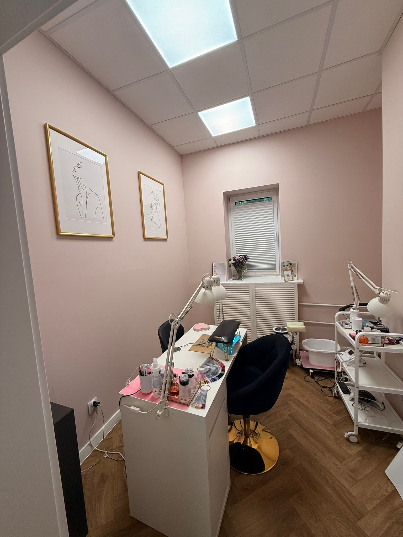 Jasny gabinet kosmetyczny z białym biurkiem, lampą i czarnym krzesłem na złotym postumencie. Na ścianach grafiki w złotych ramach. W tle okno z żaluzjami.