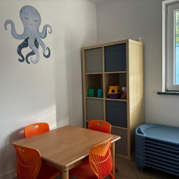 Kolorowe Rybki Aleksandra Chmielewska - Jasne wnętrze sali zabaw z drewnianym stolikiem i pomarańczowymi krzesłami. Na ścianie naklejka z ośmiornicą. Regał z zabawkami i okno z widokiem na zieleń.