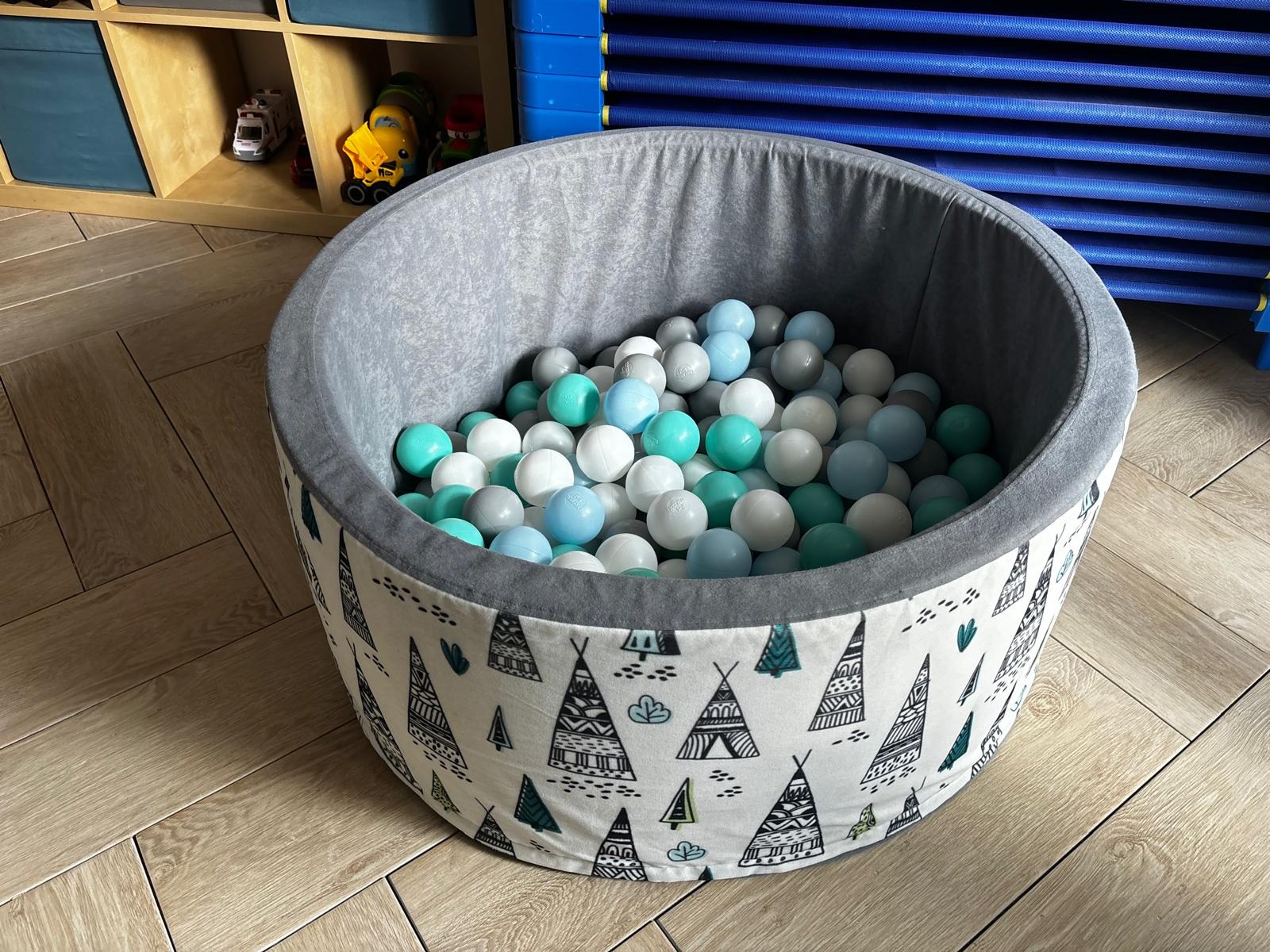 Suchy basen z kulkami w odcieniach bieli, szarości i turkusu, zdobiony wzorem tipi. Zabawka w pokoju dziecięcym z drewnianą podłogą i regałem w tle.