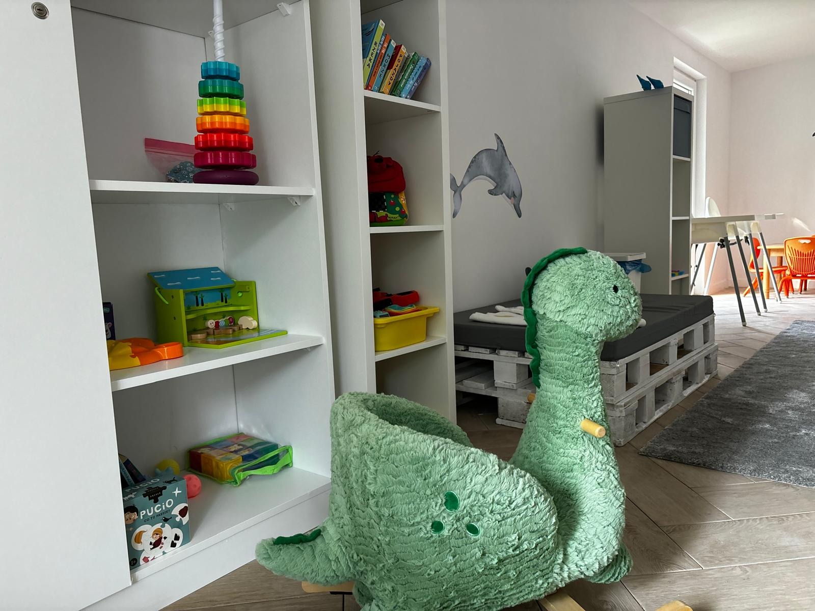 Pokój dziecięcy z białymi regałami pełnymi zabawek, miękkim dinozaurem na biegunach i naklejką delfina na ścianie, w jasnym, przestronnym wnętrzu.