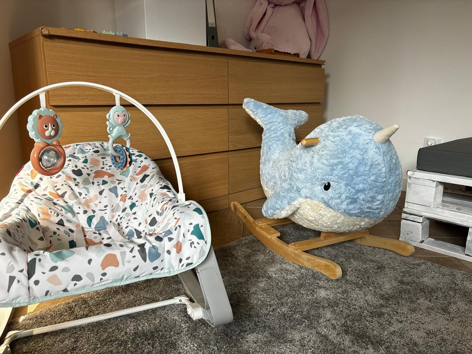 Dziecięcy bujaczek z zabawkami i pluszowy wieloryb na drewnianych płozach w pokoju dziecięcym, na tle komody i pluszowego królika.