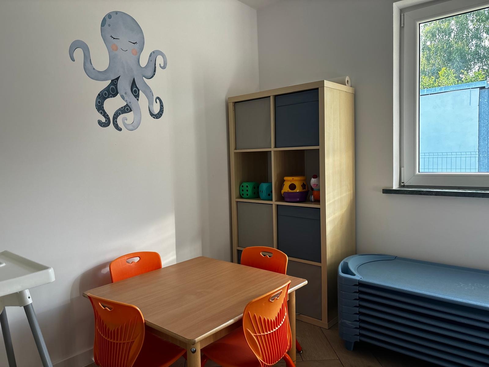 Jasne wnętrze sali zabaw z drewnianym stolikiem i pomarańczowymi krzesłami. Na ścianie naklejka z ośmiornicą. Regał z zabawkami i okno z widokiem na zieleń.