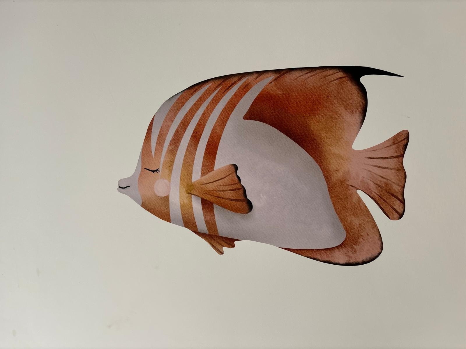 Ilustracja uśmiechniętej rybki w odcieniach brązu i bieli, z zamkniętymi oczami i delikatnym uśmiechem, umieszczona na jasnym, neutralnym tle.