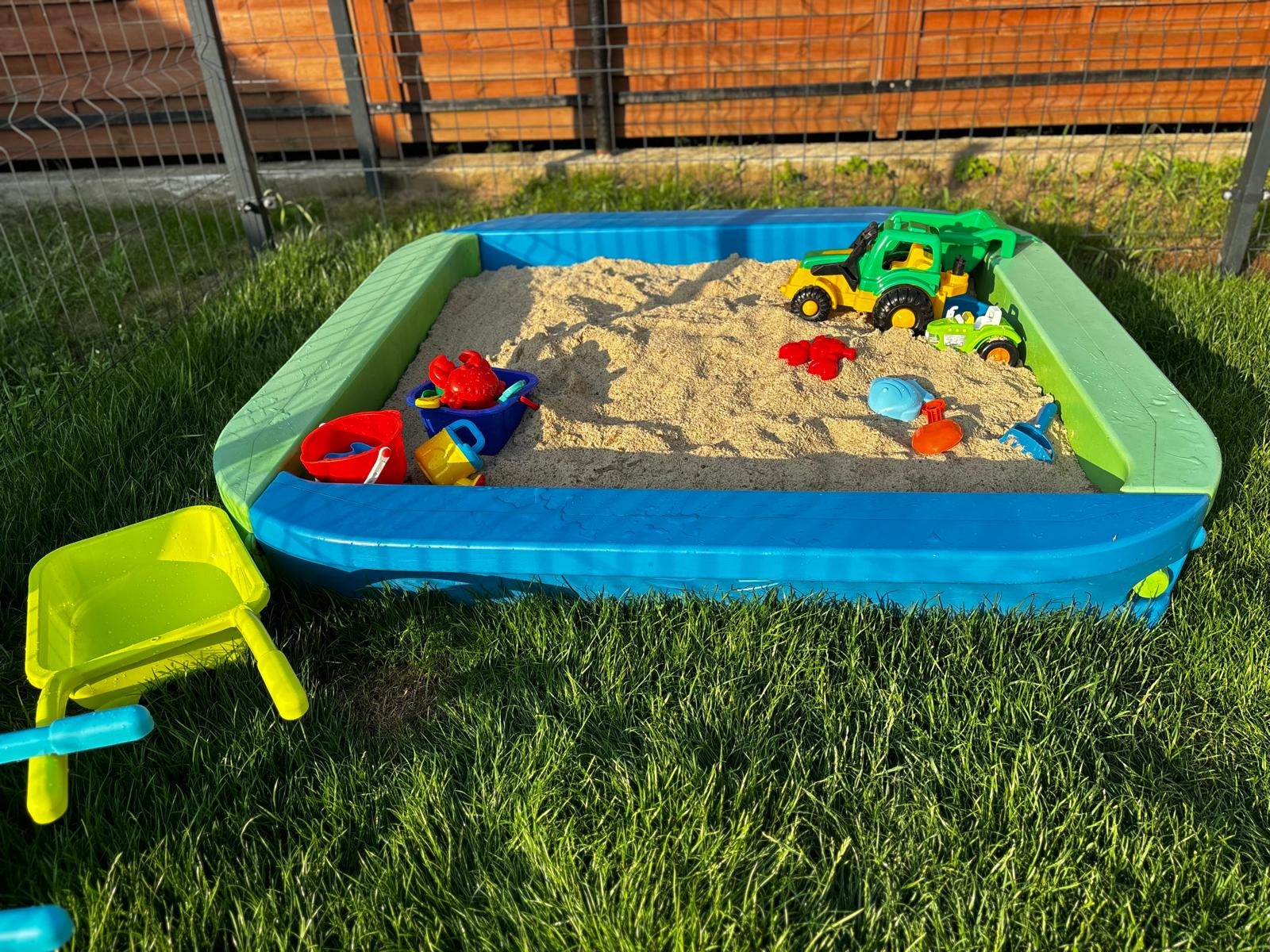 Kolorowa piaskownica z zabawkami na trawie w słoneczny dzień. Widoczne wiaderka, łopatki i plastikowe pojazdy. Idealne miejsce do zabawy na świeżym powietrzu dla dzieci.