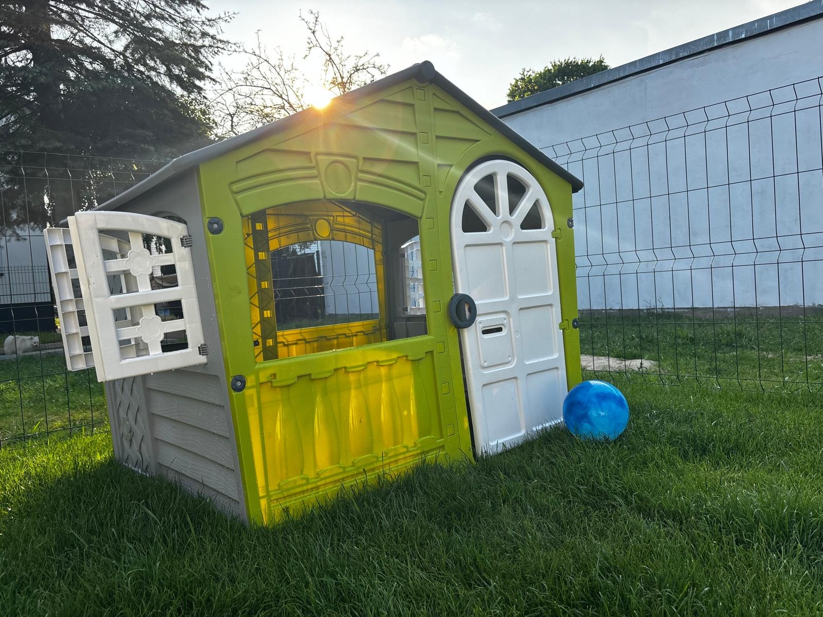 Kolorowy, plastikowy domek dla dzieci z otwartymi okiennicami i niebieską piłką na trawie, ogrodzony siatką. Słońce w tle.