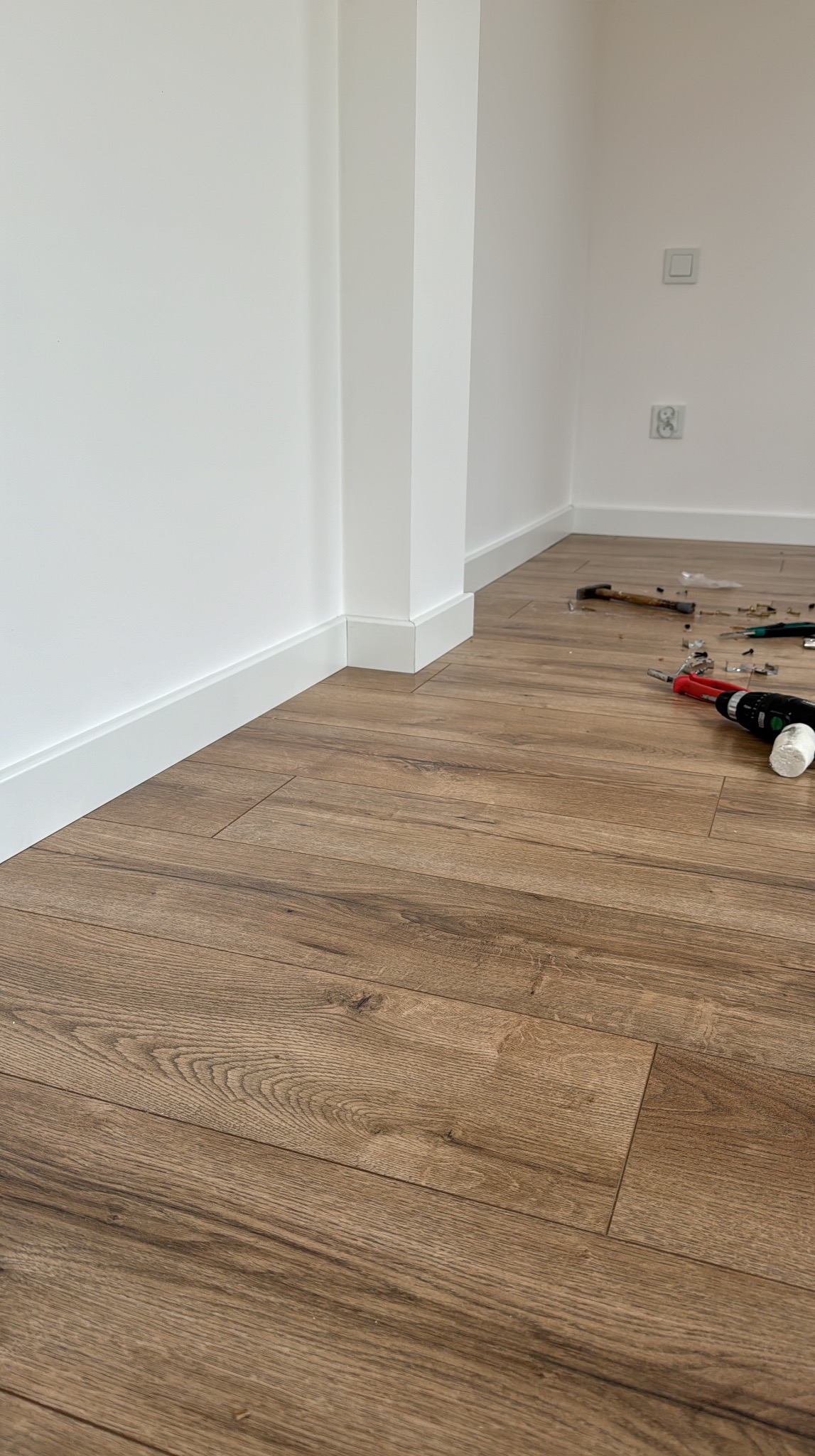 Ukończony montaż białych listew przypodłogowych MDF w rogu pomieszczenia z brązową podłogą. Widoczne narzędzia pracy i gniazdka elektryczne na ścianie.