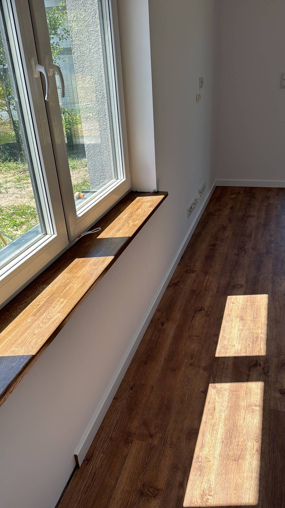 Zamontowany drewniany parapet i ułożone panele winylowe w nowym pokoju. Widoczne listwy przypodłogowe i okno z widokiem na zieleń. Słoneczne światło podkreśla strukturę podłogi.