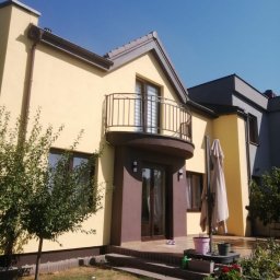 Krystian Matczak - Dwukondygnacyjny dom z balkonem, w odcieniach beżu i brązu, z elementami szarości. Widoczne rośliny w ogrodzie i błękitne niebo w tle. Słoneczny dzień.