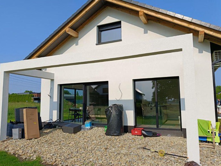 Biała elewacja domu z pergolą w trakcie wykończenia, widoczne narzędzia i materiały budowlane, jasne tło, słoneczny dzień.