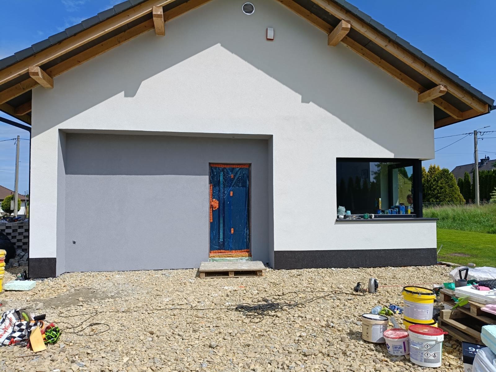 Dwukolorowa elewacja domu z oknem i zabezpieczonymi drzwiami, widoczne wiadra z farbą i narzędzia na żwirowym podłożu. Jasne niebo w tle.