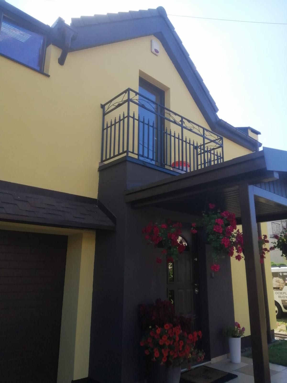 Żółto-brązowy dom jednorodzinny z balkonem o stalowej balustradzie i kwiatami w donicach, elewacja po renowacji. Widok z dołu, słoneczny dzień.