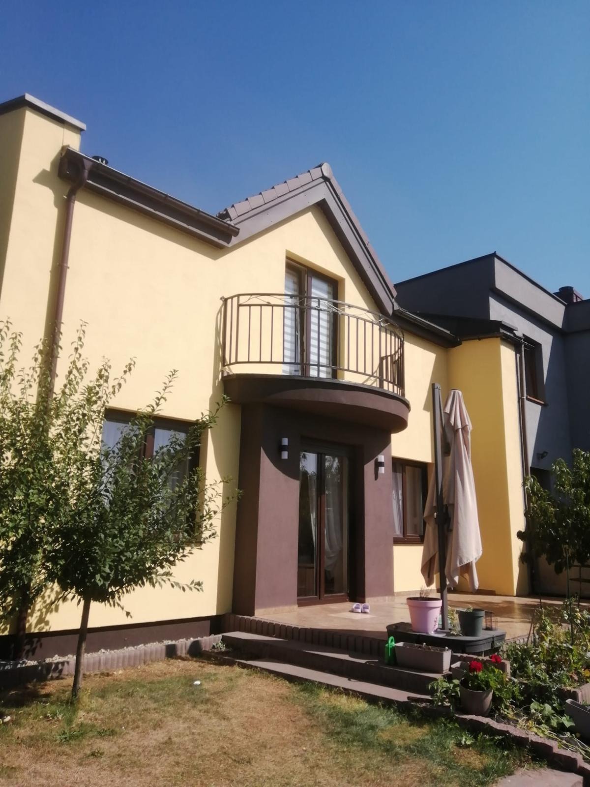 Dwukondygnacyjny dom z balkonem, w odcieniach beżu i brązu, z elementami szarości. Widoczne rośliny w ogrodzie i błękitne niebo w tle. Słoneczny dzień.