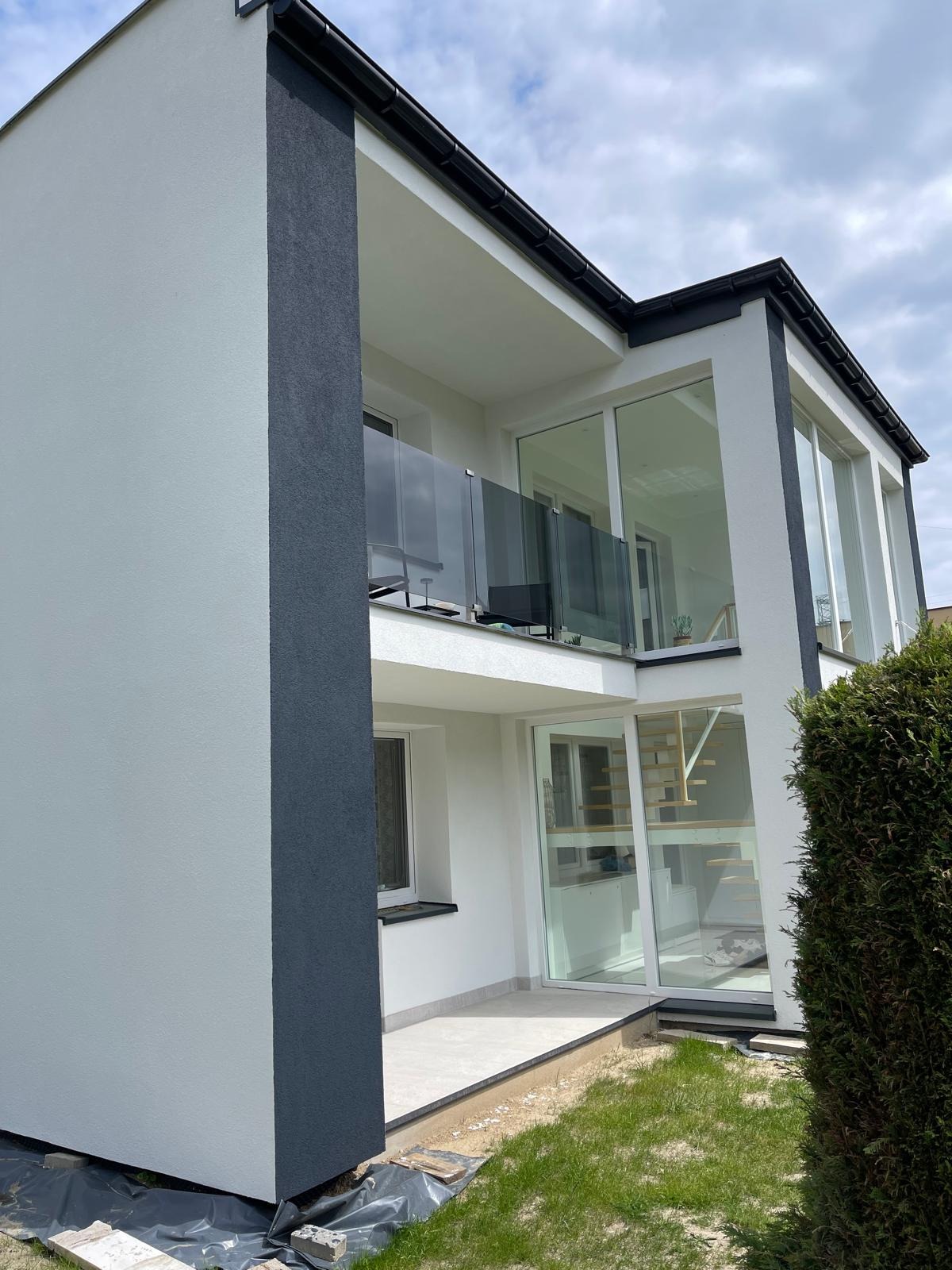 Nowoczesny dom z jasną elewacją i ciemnymi akcentami, przeszklony balkon z szarymi balustradami, widok z zewnątrz, trawnik w pierwszym planie.