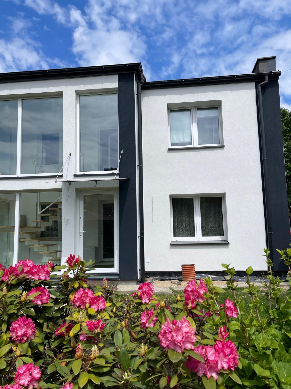 Nowoczesny dom z białą elewacją i grafitowymi akcentami, otoczony kwitnącymi różanecznikami. Widoczne duże okna i przeszklone wejście.