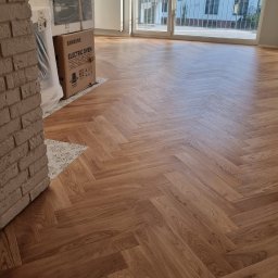 MPodłogi - Ułożona jodełka francuska z drewnianych paneli w jasnym wnętrzu z dużym oknem balkonowym. Częściowo widoczny ceglany filar i kartony z AGD.