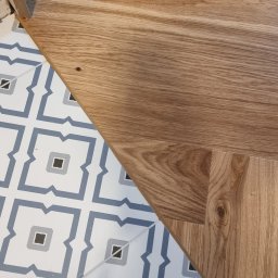 MPodłogi - Fragment schodów z jasnego drewna i przylegającej podłogi z płytek w biało-niebieskie wzory. Widoczny detal wykończenia i kontrast materiałów.