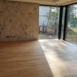 MPodłogi - Jasne, drewniane deski podłogowe w przestronnym wnętrzu z dużymi oknami, wpadające promienie słoneczne tworzą grę światła i cienia. Widoczna ściana z marmurowym wzorem.