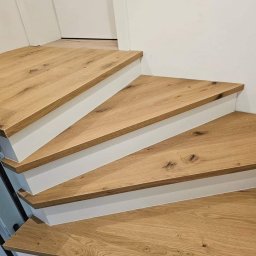 MPodłogi - Drewniane schody o jasnym odcieniu, z białymi podstopnicami, ukazane z bliska. Widoczne słoje drewna i detale wykończenia. Minimalistyczny design.