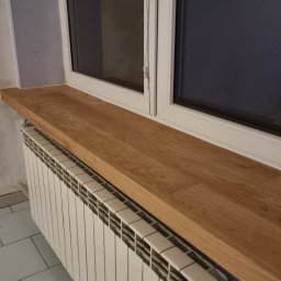 MPodłogi - Jasny, dębowy parapet z widocznym usłojeniem, zamontowany nad grzejnikiem w pokoju z białymi oknami i szarą ścianą. Widok z dołu, ukośny.