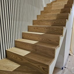 MPodłogi - Drewniane schody o jasnym odcieniu dębu, z widocznym rysunkiem słojów, kontrastują z białą ścianą dekorowaną pionowymi listwami. Wykończenie w trakcie prac.