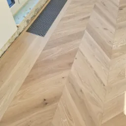 Jasny parkiet ułożony we wzór jodełki francuskiej, z widoczną kratką wentylacyjną przy oknie. Wykończona podłoga z naturalnego drewna.