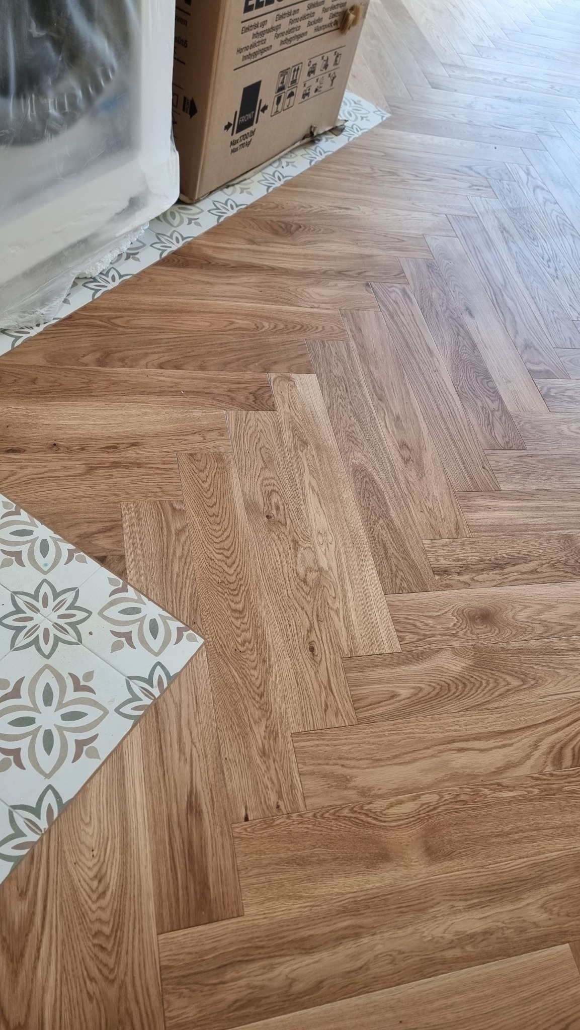 Ułożona podłoga z paneli w jodełkę, widoczny fragment płytki podłogowej z wzorem kwiatowym i karton. Ciepłe odcienie drewna i subtelny wzór tworzą elegancki efekt.