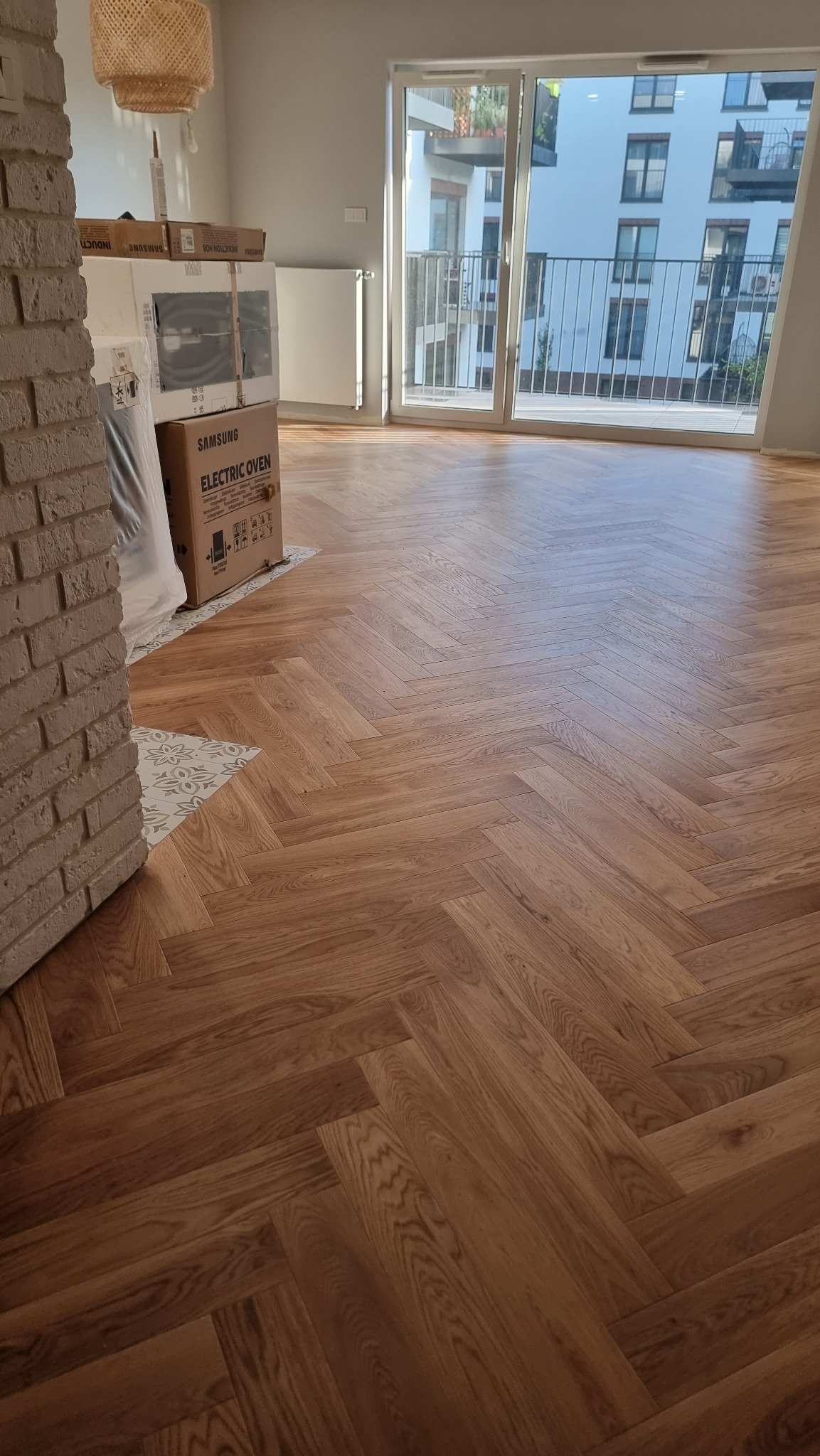 Ułożona jodełka francuska z drewnianych paneli w jasnym wnętrzu z dużym oknem balkonowym. Częściowo widoczny ceglany filar i kartony z AGD.