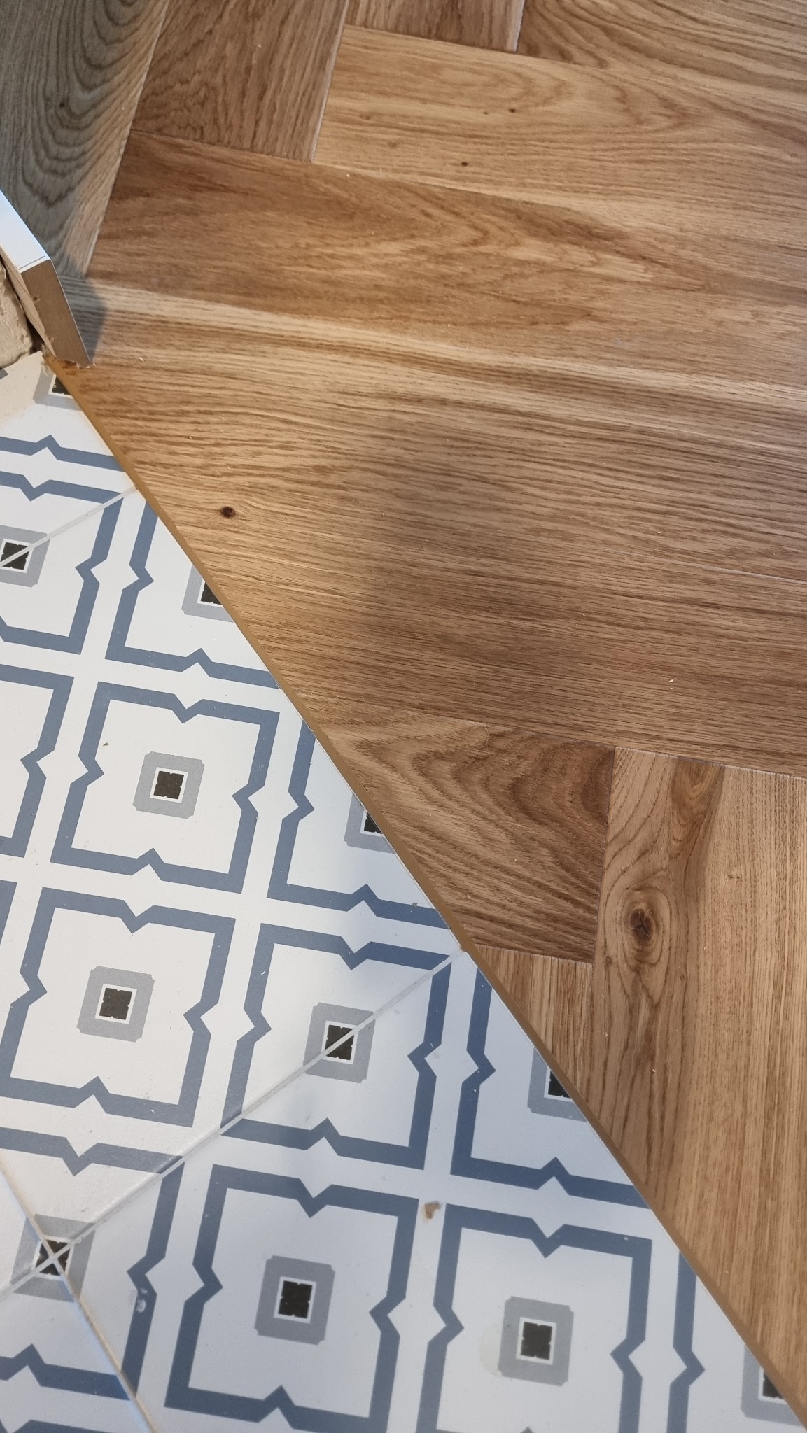 Fragment schodów z jasnego drewna i przylegającej podłogi z płytek w biało-niebieskie wzory. Widoczny detal wykończenia i kontrast materiałów.