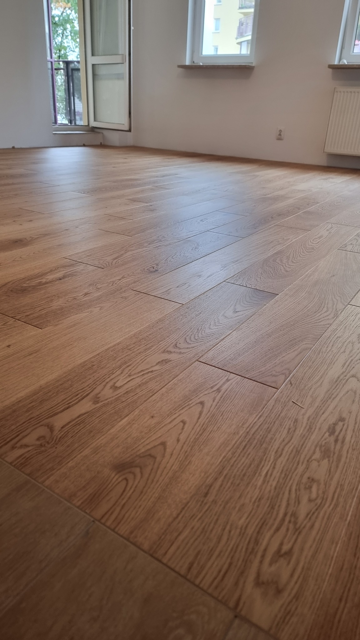 Ułożony parkiet w jasnym odcieniu drewna w nowym mieszkaniu. Widoczne okno i drzwi balkonowe. Podłoga błyszczy w świetle.