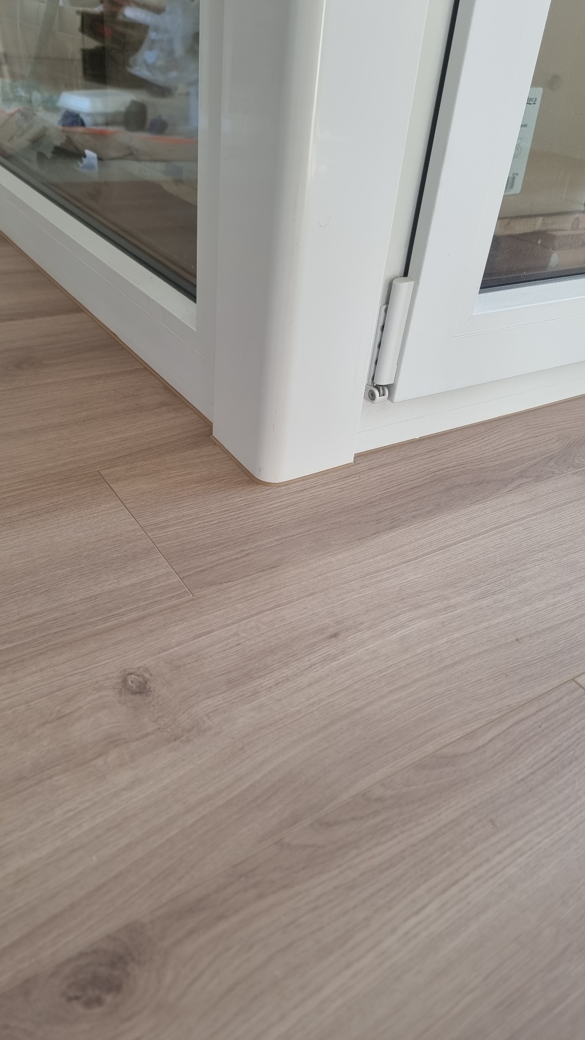 Jasne panele podłogowe ułożone przy białej ramie okiennej. Widoczne subtelne słoje drewna i delikatna fuga między panelami. Detal wykończenia wnętrza.