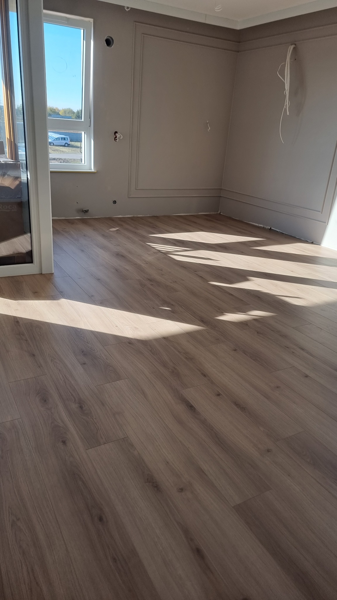 Jasne panele podłogowe w nowym wnętrzu, z promieniami słońca i dekoracyjną listwą przypodłogową. Widok przez okno na zewnątrz.