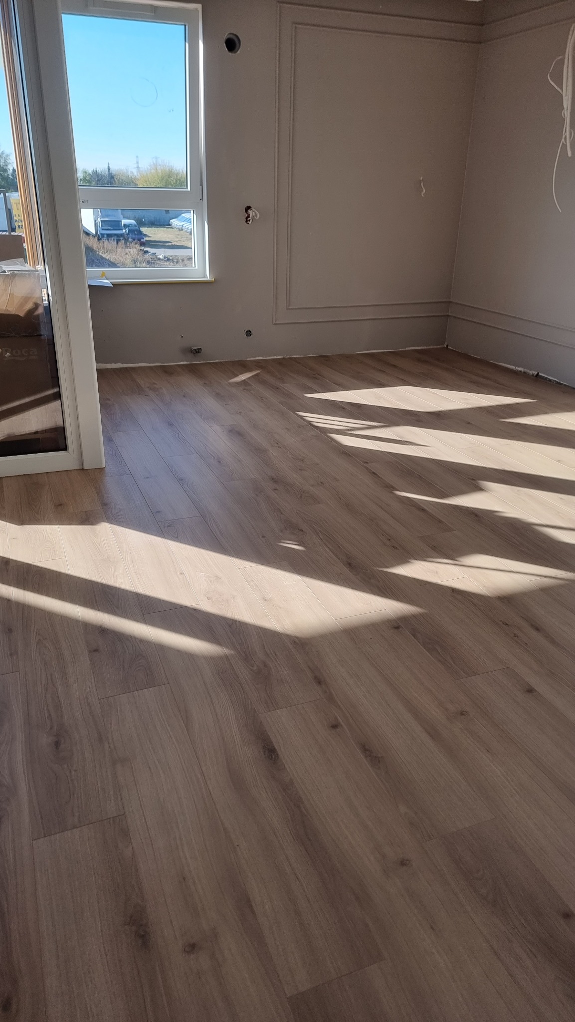 Jasne panele podłogowe w nowym wnętrzu, oświetlone promieniami słońca padającymi przez okno. Ściany w kolorze beżowym z dekoracyjnymi listwami.