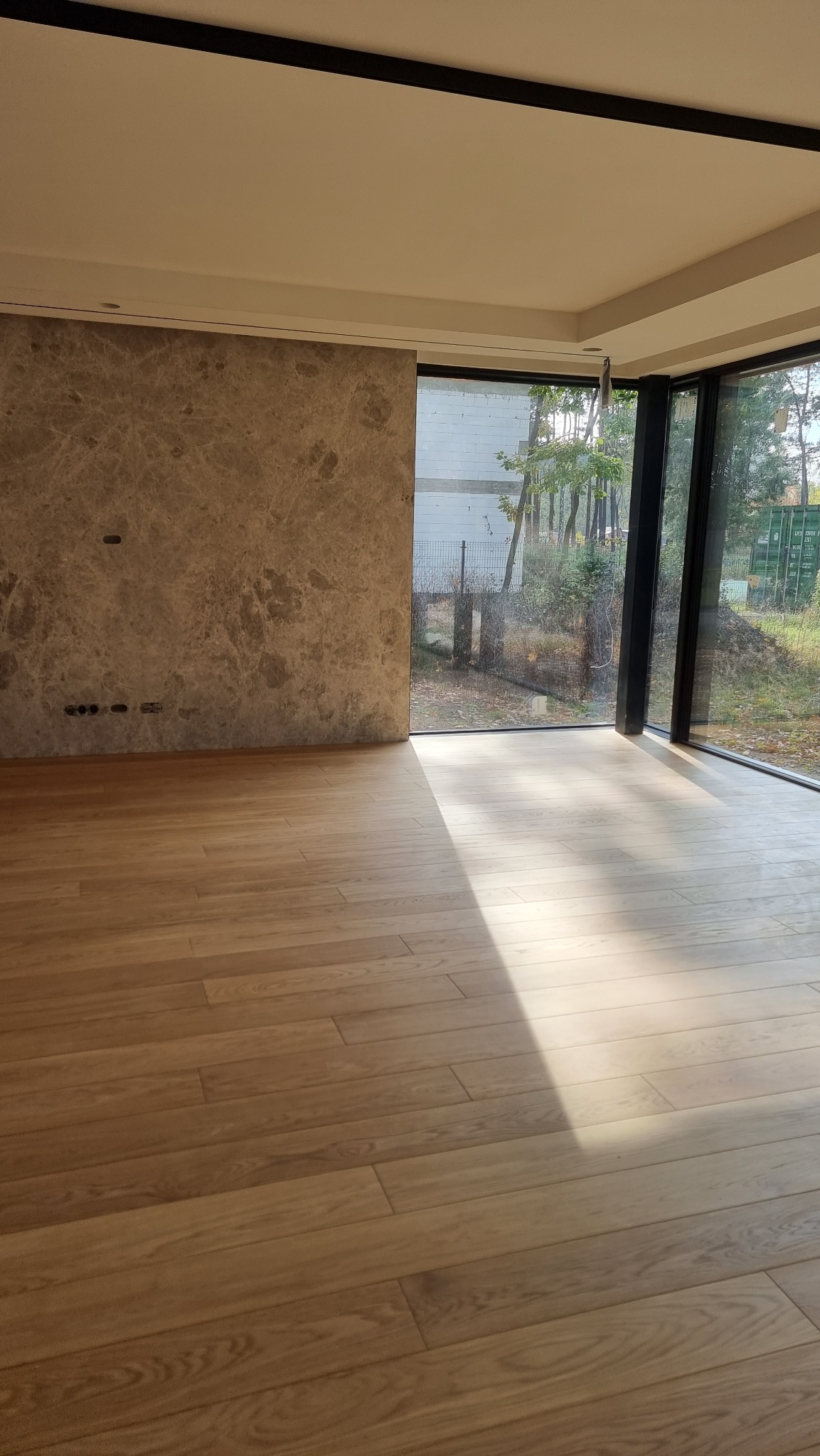 Jasne, drewniane deski podłogowe w przestronnym wnętrzu z dużymi oknami, wpadające promienie słoneczne tworzą grę światła i cienia. Widoczna ściana z marmurowym wzorem.